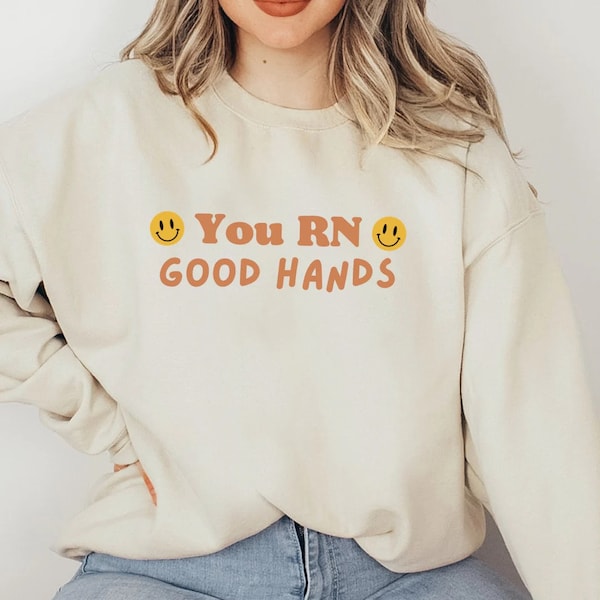May include: A cream colored sweatshirt with the text "You RN Good Hands" in orange and brown lettering. Two yellow smiley faces are on either side of the text.