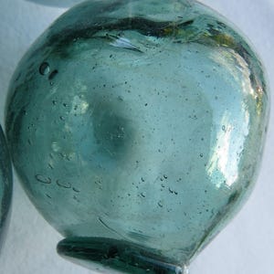 May include: A round, translucent, light blue-green glass float with a slightly flattened base. The surface of the glass is textured with numerous small air bubbles and subtle imperfections, creating a reflective and organic appearance.