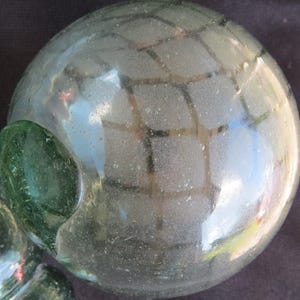May include: A clear, round glass float with a green glass attachment. The float has a grid-like pattern on its surface and is slightly frosted. The glass has a reflective quality, with hints of blue and green visible.