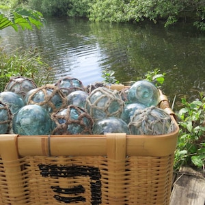 May include: A wicker basket filled with a collection of glass fishing floats, some with netting, in shades of blue and green. The basket is sitting on a wooden jetty near a pond.