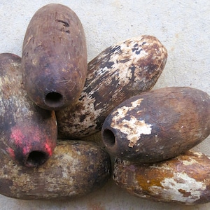 May include: A collection of six weathered, oval-shaped wooden fishing floats. Each float features a central hole, with surfaces showing signs of wear, including patches of peeling white paint, dark brown wood grain, and a small splash of red paint.
