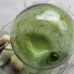 May include: A spherical, translucent light green glass float with subtle internal swirls and a small, darker green glass seal. The glass orb is positioned alongside several off-white, spiral-shaped seashells and thin, light-colored netting.
