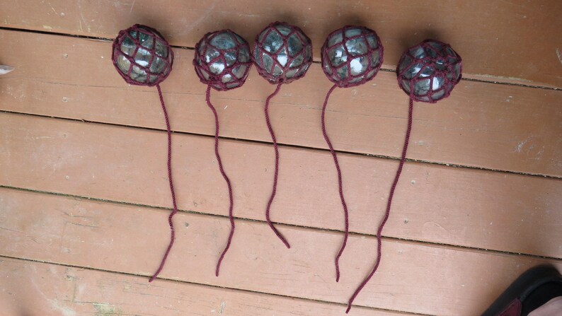 May include: Six glass floats with burgundy netting and strings hanging from a wooden surface.