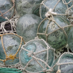 May include: A collection of glass fishing floats, some with a rope net design, in various shades of blue and green. The floats are arranged on a bed of white and gray netting.