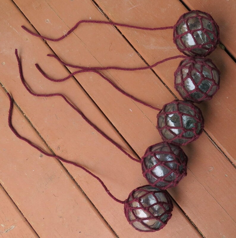 May include: Five glass floats with a dark brown net covering each one. The floats are hanging from maroon cords.