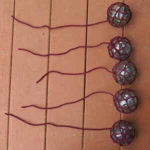 May include: Five glass floats with a dark red net covering each one. The floats are hanging from dark red strings.