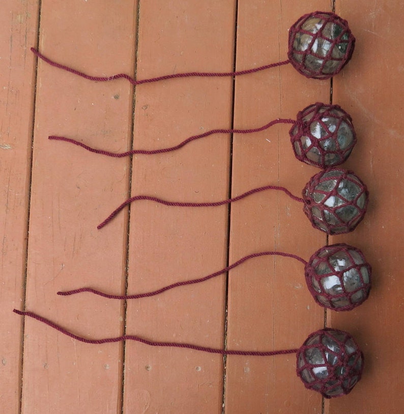 May include: Five glass floats with a dark red net covering each one. The floats are hanging from dark red strings.