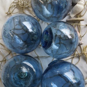 Puede incluir: Cinco boyas de pesca de vidrio azul con redes oscuras en el interior, dispuestas sobre una red beige. Los flotadores tienen un acabado brillante y diferentes tonos de azul. También se ven conchas marinas y un trozo de madera flotante.