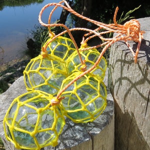 Op de afbeelding: Vier heldere glazen visdrijvers met geel net en oranje touw zijn gerangschikt op een verweerde houten paal. De drijvers hebben een lichte groene tint en zijn samengebonden met oranje touw.