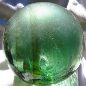 May include: A clear, green glass gazing ball with a spherical shape. The glass has a slightly textured surface with dark green streaks and small air bubbles. The ball reflects light, creating highlights and shadows.