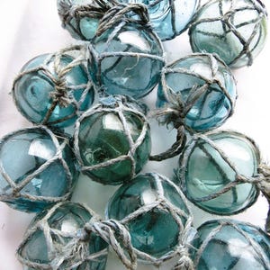 May include: A collection of clear, blue-green glass fishing floats, each encased in a knotted rope netting. The floats vary slightly in color and size, creating a textured and visually interesting display. The nautical theme is evident.