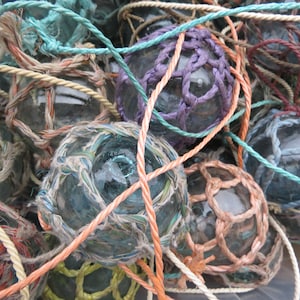 May include: A close-up of a pile of glass fishing floats, each wrapped in colourful rope. The floats are various shades of blue, green, and purple, and the ropes are a mix of white, orange, and blue.