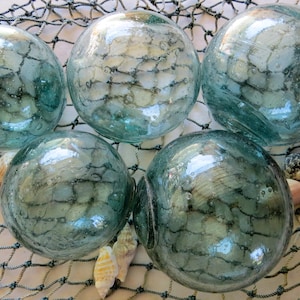 May include: Five spherical, translucent, light blue-green glass floats arranged on a dark, knotted fishing net. Each glass orb features visible air bubbles within the material, and several small, light-colored seashells are scattered among the net.
