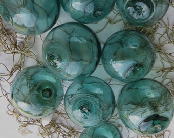 May include: A collection of nine teal-colored glass fishing floats. The spheres vary slightly in size and feature internal marbling. They rest on a beige fishing net, creating a nautical theme. The glass has a reflective quality.