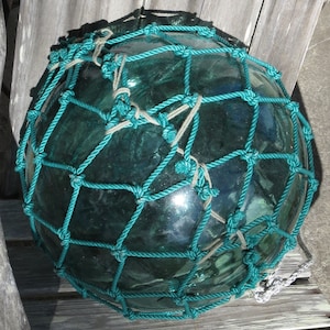 May include: A large, round, green glass buoy with a teal blue rope net. The buoy is used for fishing and is a popular nautical decor item.