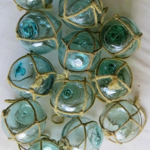 May include: A collection of eleven spherical glass fishing floats in varying shades of light blue and seafoam green. Each translucent glass orb is encased in a decorative, hand-tied tan jute rope netting, featuring a central circular seal mark.
