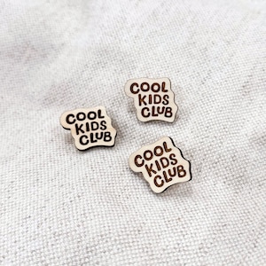 May include: Three wooden pins with the text "Cool Kids Club" in a wavy font. The pins are light brown and the text is dark brown.