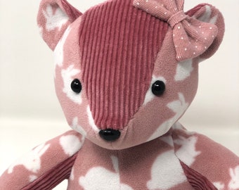 Peut inclure: Un ours en peluche rose avec un motif de lapin blanc et un nœud rose à pois blancs. L'ours a des yeux noirs et un nez noir.