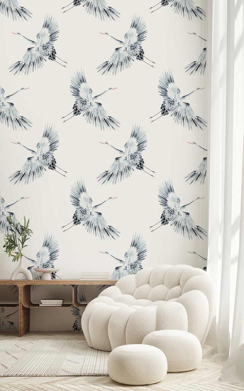 May include: A white room with a white couch and two white ottomans. The wall is covered in a wallpaper with a pattern of blue cranes.