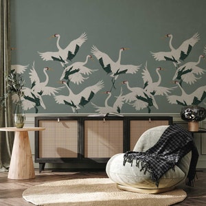 May include: A living room interior with a light green wall featuring a wallpaper pattern of white cranes flying in a flock. The room has a wooden side table, a black cabinet with woven rattan doors, and a light gray armchair with a black and white throw blanket.