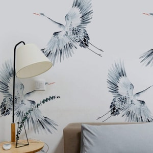 May include: A bedroom with a white wall featuring a repeating pattern of gray watercolor cranes. The bed has a gray duvet and two gray pillows. A wooden side table with a lamp and a vase with greenery is next to the bed.