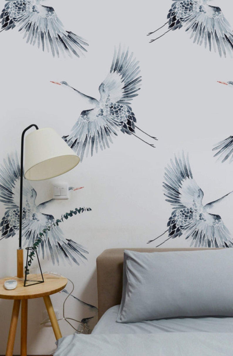 May include: A bedroom with a white wall featuring a repeating pattern of gray watercolor cranes. The bed has a gray duvet and two gray pillows. A wooden side table with a lamp and a vase with greenery is next to the bed.
