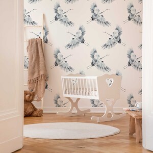 May include: A white wooden rocking cradle with a heart-shaped cutout on the headboard. The cradle is in a nursery with a white and pink wallpaper featuring a repeating pattern of blue cranes. A brown knitted blanket is draped over a wooden ladder in the background.