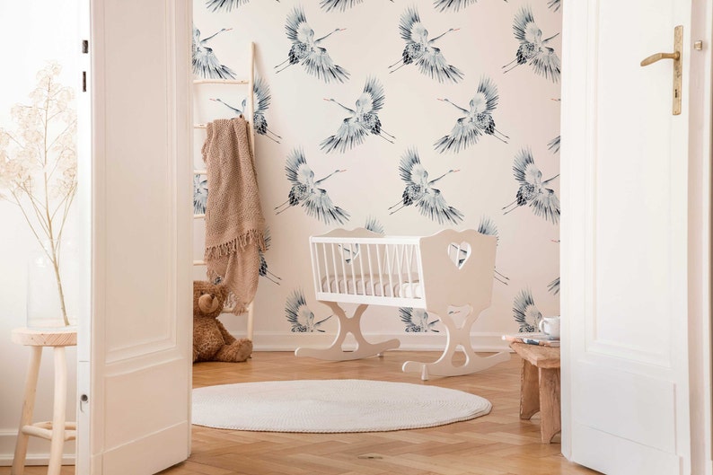 May include: A white wooden rocking cradle with a heart-shaped cutout on the headboard. The cradle is in a nursery with a white and pink wallpaper featuring a repeating pattern of blue cranes. A brown knitted blanket is draped over a wooden ladder in the background.
