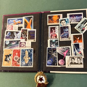 May include: A collection of vintage postage stamps and a commemorative pin. The stamps, housed in a black album, feature space exploration themes with rockets, astronauts, and celestial bodies. The pin has a red star and a ribbon.