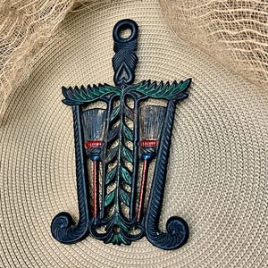 May include: A black cast iron trivet featuring a central vertical leaf motif flanked by two broom designs. The piece includes green accents on the leaves and red and blue details on the broom handles. It has a top hanging loop and scroll feet.