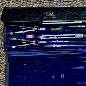 May include: A black leather case containing a set of silver metal drafting instruments. The case is labeled POST TRUEMASTER 1085 C and features a blue velvet interior. The set includes various compasses, dividers, and attachments for technical drawing.