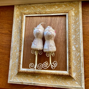 May include: Two white fabric bust forms with lace trim and pink and green embellishments are displayed on a white metal stand. The stand is hanging on a gold and white ornate picture frame.