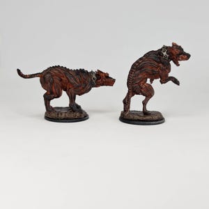 May include: Two brown and black dog figurines with spiked collars. One is in a walking pose, the other is standing on its hind legs. Both are on small, round, textured bases. The figurines have a detailed, textured appearance.