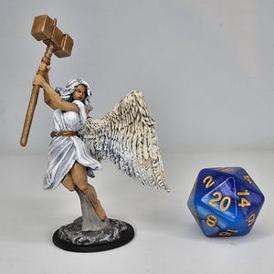 May include: A white painted miniature figurine of an angel with wings, holding a golden hammer. The angel is wearing a white robe and has long white hair. The figurine is standing on a gray base. A blue and white 20-sided die is in the background.