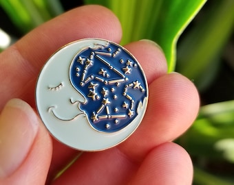 May include: A blue and white enamel pin featuring a crescent moon with a sleeping face and a constellation of stars. The pin is a whimsical and celestial design.