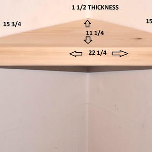 May include: A light-colored wooden corner shelf with a triangular shape. The shelf features printed dimensions: 1 1/2 thickness, 15 3/4 sides, 11 1/4 depth, and 22 1/4 front width. The shelf is mounted in a corner against a plain white wall.