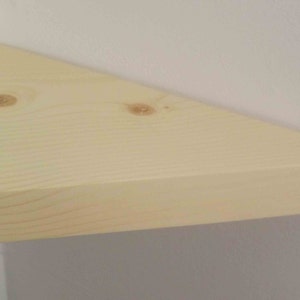 May include: A triangular corner shelf made of light-colored natural wood with visible grain patterns and two small knots on the surface. The shelf is mounted flush against two perpendicular white walls, creating a clean and simple storage surface.