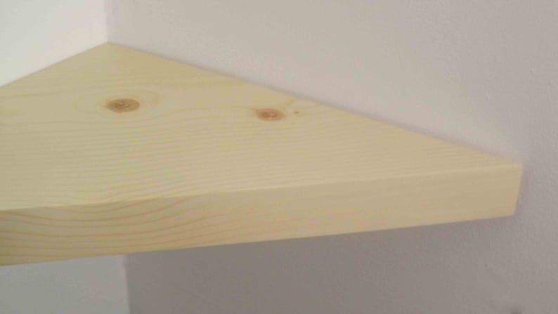 May include: A triangular corner shelf made of light-colored natural wood with visible grain patterns and two small knots on the surface. The shelf is mounted flush against two perpendicular white walls, creating a clean and simple storage surface.