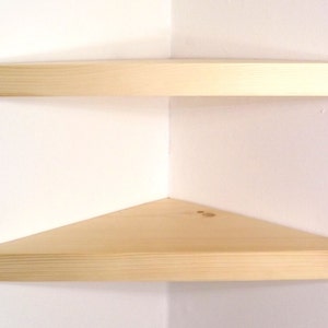 May include: Two light-colored wooden corner shelves mounted on a white wall. Each shelf features a triangular shape with a smooth, natural wood grain finish, designed to fit flush into the corner of a room for storage or display purposes.