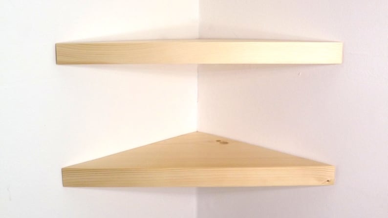 May include: Two light-colored wooden corner shelves mounted on a white wall. Each shelf features a triangular shape with a smooth, natural wood grain finish, designed to fit flush into the corner of a room for storage or display purposes.