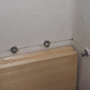 May include: A light-colored wooden board is mounted against a white wall using several silver-toned metal screws. The screws are spaced along a horizontal line where the wooden board meets the wall, with one screw visible on the adjacent wall section.