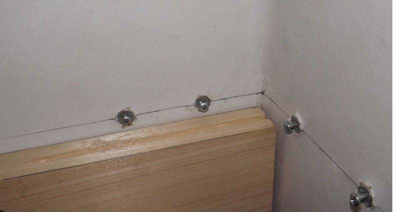 May include: A light-colored wooden board is mounted against a white wall using several silver-toned metal screws. The screws are spaced along a horizontal line where the wooden board meets the wall, with one screw visible on the adjacent wall section.
