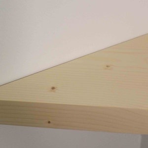 May include: A triangular corner shelf made of light-colored natural wood with visible grain patterns and small knots. The shelf is mounted flush against two perpendicular white walls, creating a clean, functional storage surface in the corner.