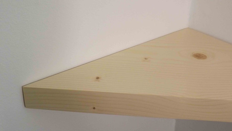 May include: A triangular corner shelf made of light-colored natural wood with visible grain patterns and small knots. The shelf is mounted flush against two perpendicular white walls, creating a clean, functional storage surface in the corner.