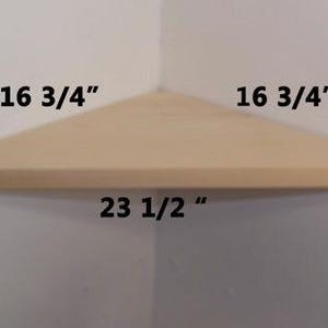 May include: A light-colored wooden corner shelf with a triangular shape. The dimensions are printed on the image as 16 3/4 inches for the two sides that meet the wall and 23 1/2 inches for the front edge facing outward from the corner.