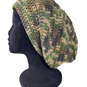 Peut inclure: Un bonnet au crochet à motif camouflage. Le chapeau présente un mélange de fils verts, bruns et gris, créant un aspect texturé. Le chapeau est présenté sur une tête de mannequin noire, mettant en valeur sa forme et son design. Ce chapeau fait main est adapté au froid.