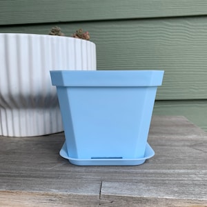 May include: A light blue plastic square planter with a matching saucer. The planter has a slightly textured surface.