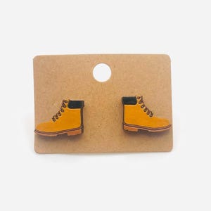 May include: A pair of yellow and black boot-shaped earrings. The earrings are attached to a brown card with a circular hole at the top. The boots have black collars and brown soles. The earrings are a miniature version of work boots.