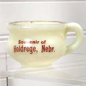 May include: A small, off-white souvenir cup with a handle. The cup has the words "Souvenir of Holdrege, Nebr." in red lettering. The cup is sitting on a clear, glass shelf.