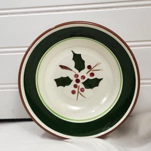 May include: A round ceramic bowl with a white interior featuring a holly and berry design. The bowl has a dark green band, a light green accent, and a brown rim. The bowl is likely for serving food.
